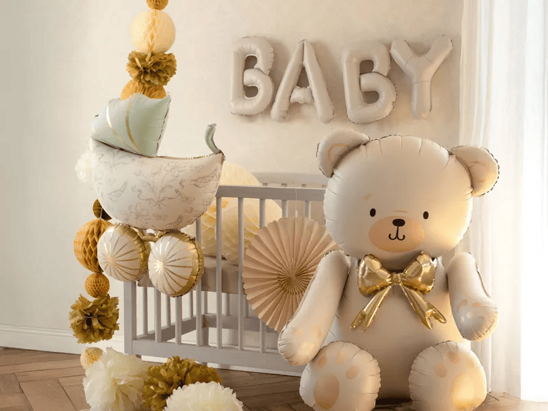 Allestimento festa nascita neutro con palloncini Baby oro e beige, orsetto gigante in foil, carrozzina decorativa e scritta Baby su sfondo con culla bianca.