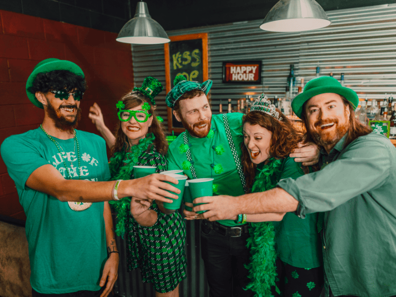 Festa di San Patrizio in pub: amici con outfit verdi a tema Irlanda brindano con bicchieri, atmosfera St. Patrick’s Day